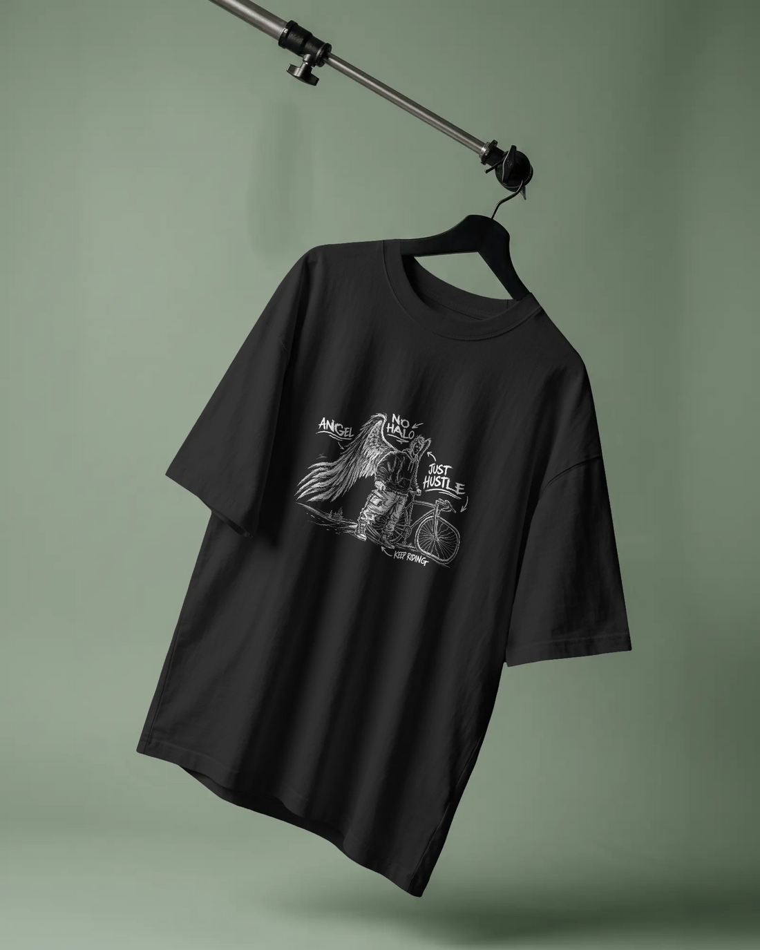 Black Cycle Swagger No Halo Just Hustle T-shirt featuring a white chalk-style winged cyclist illustration, hanging against a muted green studio background.