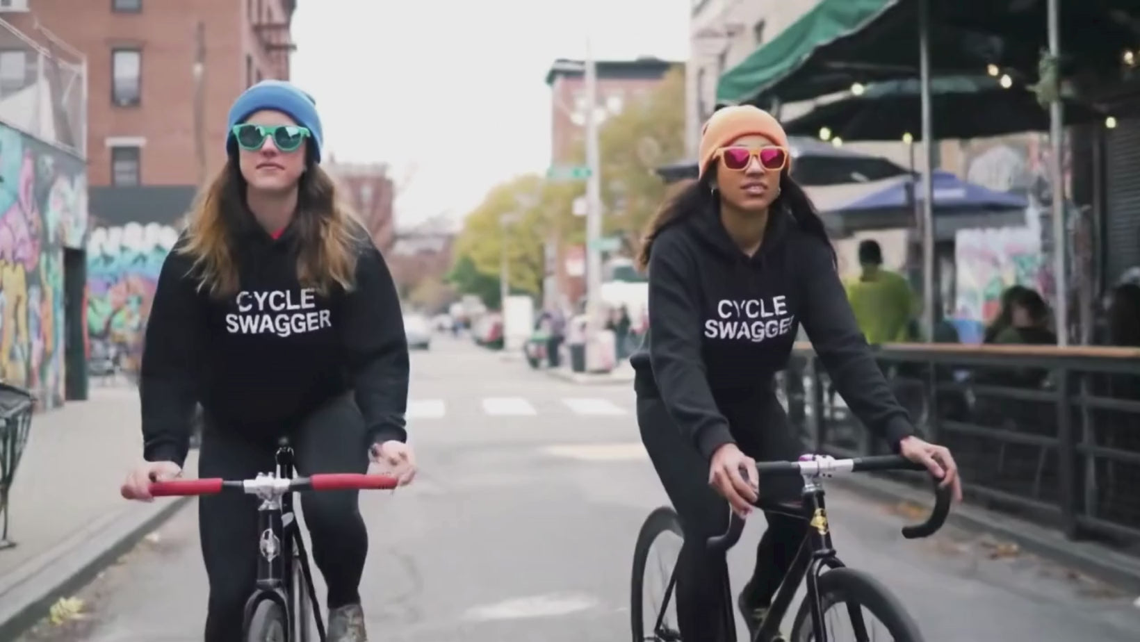 Load video: Two cyclists wearing black Cycle Swagger hoodies, beanies, and sunglasses, ride bikes side-by-side on an urban street with graffiti.